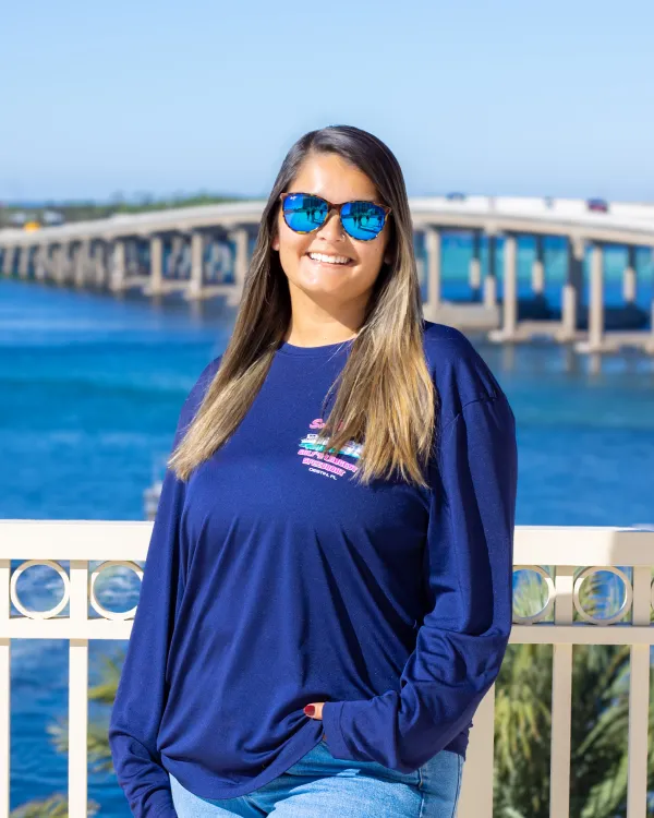 Person in sunglasses and blue shirt stands by a railing with a bridge over water in the background.