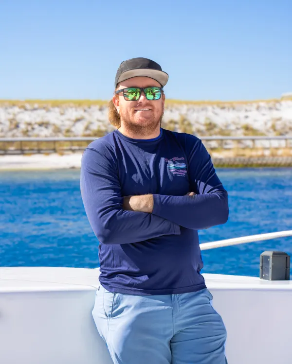 Man in sunglasses and cap standing on boat with arms crossed.