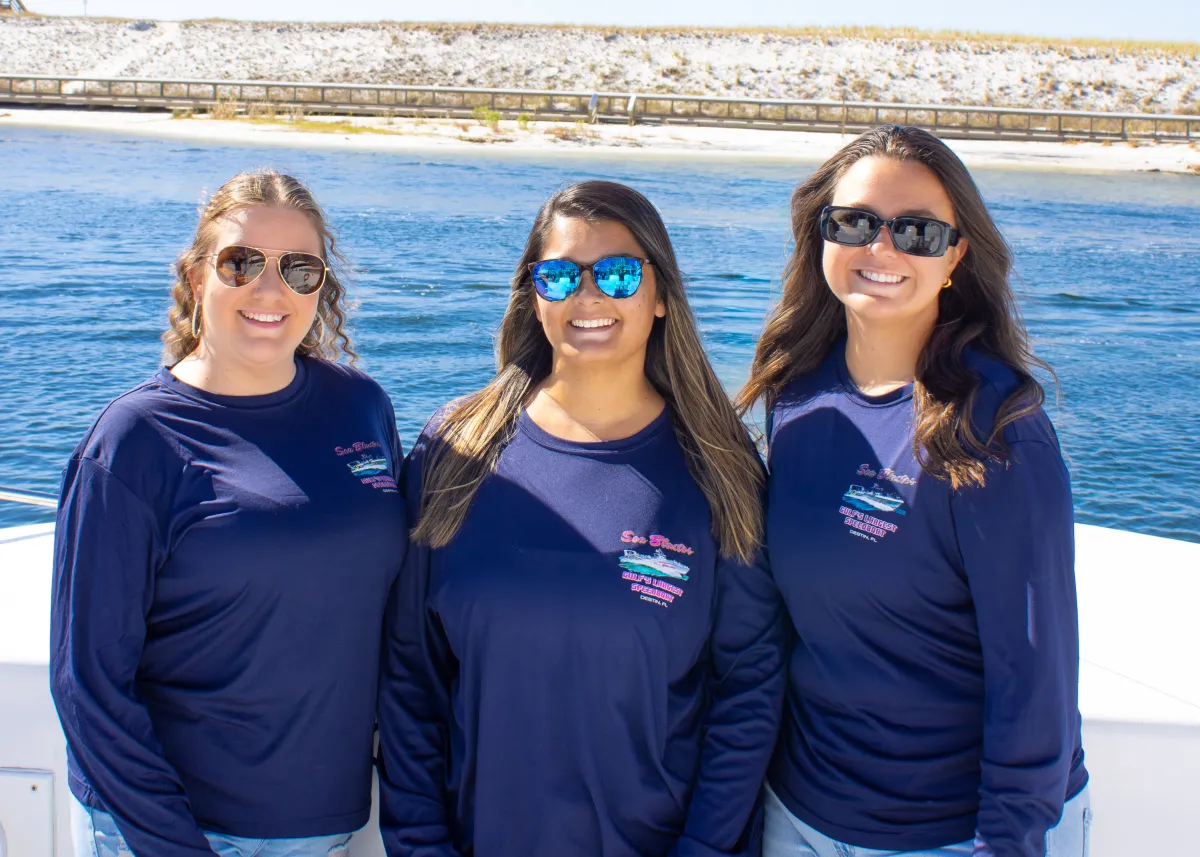 Three women in sunglasses and blue shirts standing on a boat near water.
