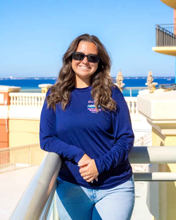 Woman in sunglasses leaning on railing, smiling, with sea and buildings in background.