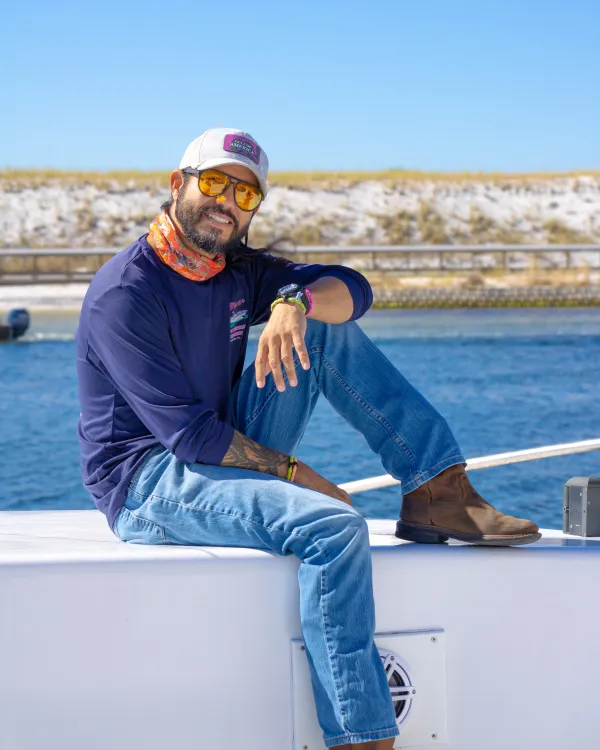 Smiling man in casual attire sits on boat deck with blue sky background.