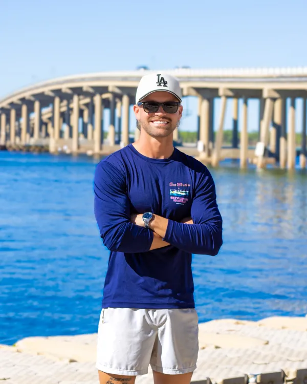 Smiling man in cap and sunglasses stands by a river with a bridge in the background.