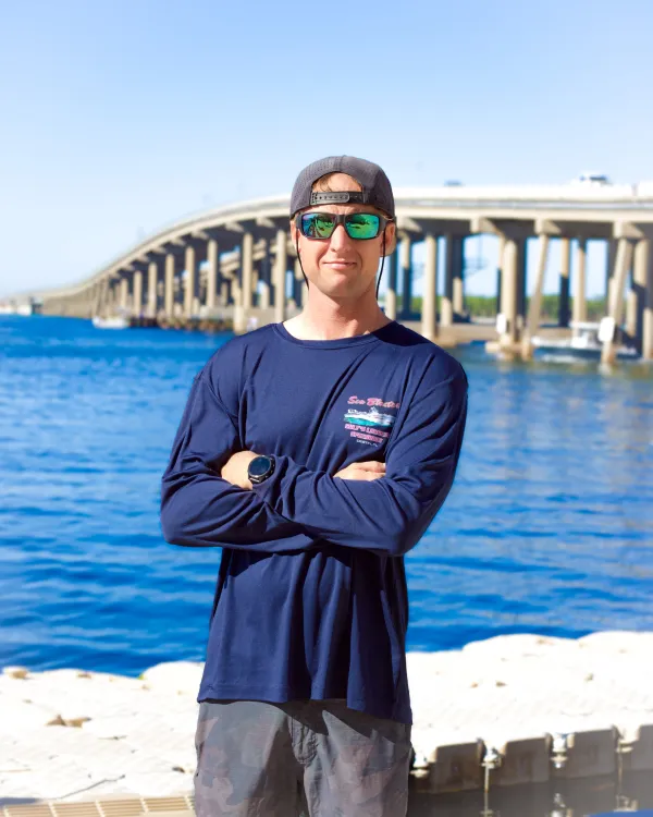 Man in sunglasses and cap stands by water with bridge in background.