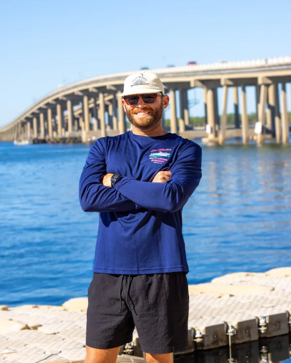 Person in blue shirt and hat standing by a waterfront with a bridge in the background.